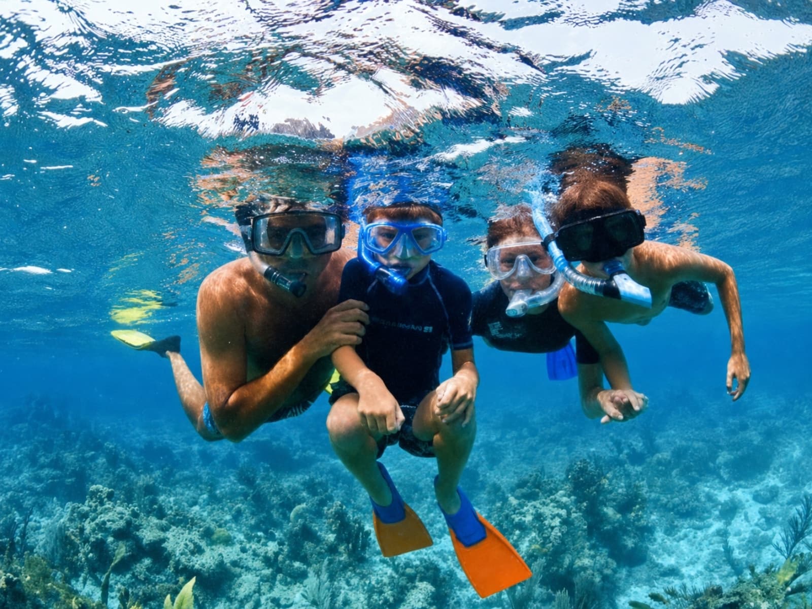 A family in snorkel masks swims toward the camera under the turquoise water, above the reef