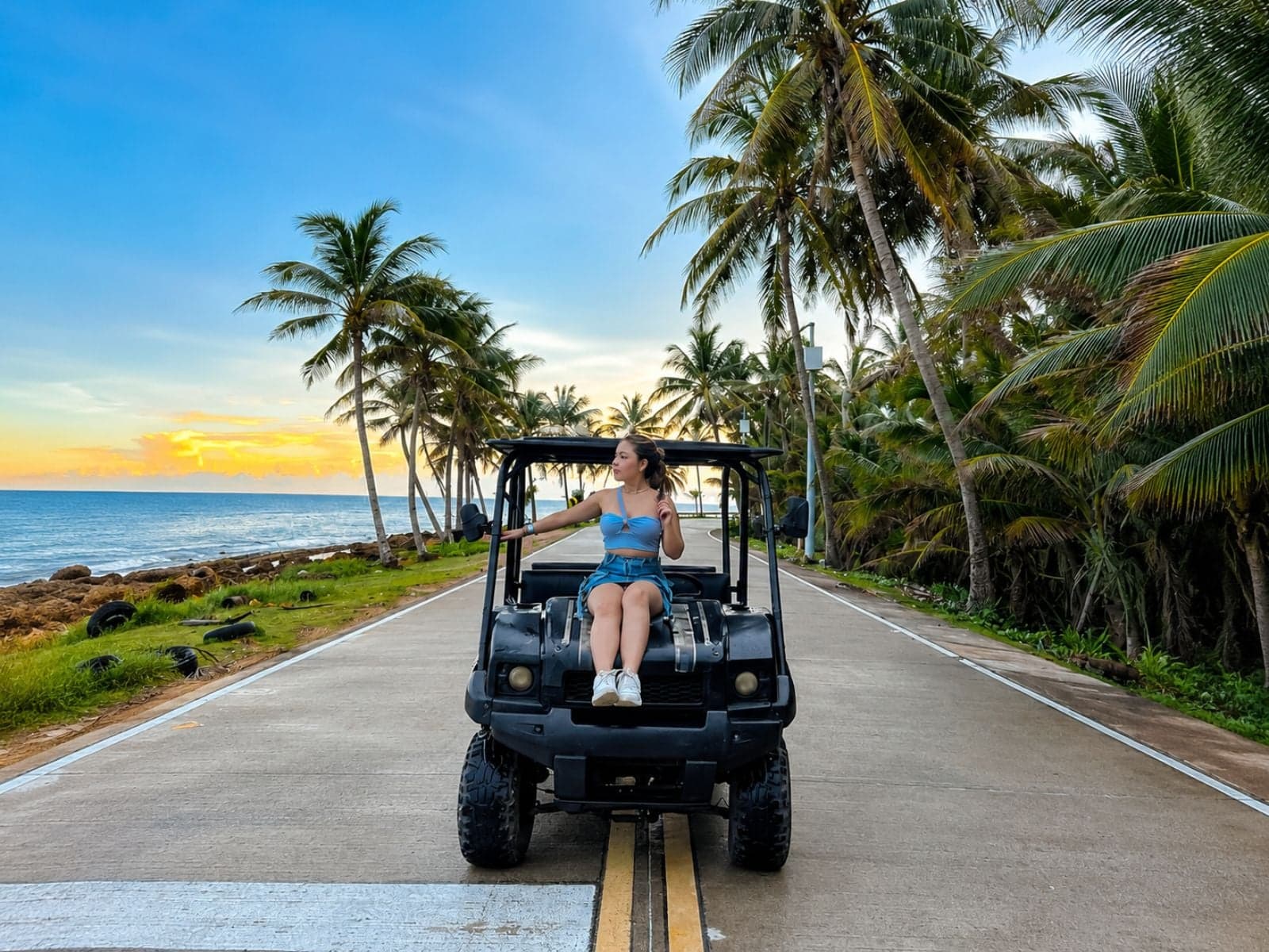 Traveler sitting in a golf cart on the ring road, among palm trees, with the sea at sunset