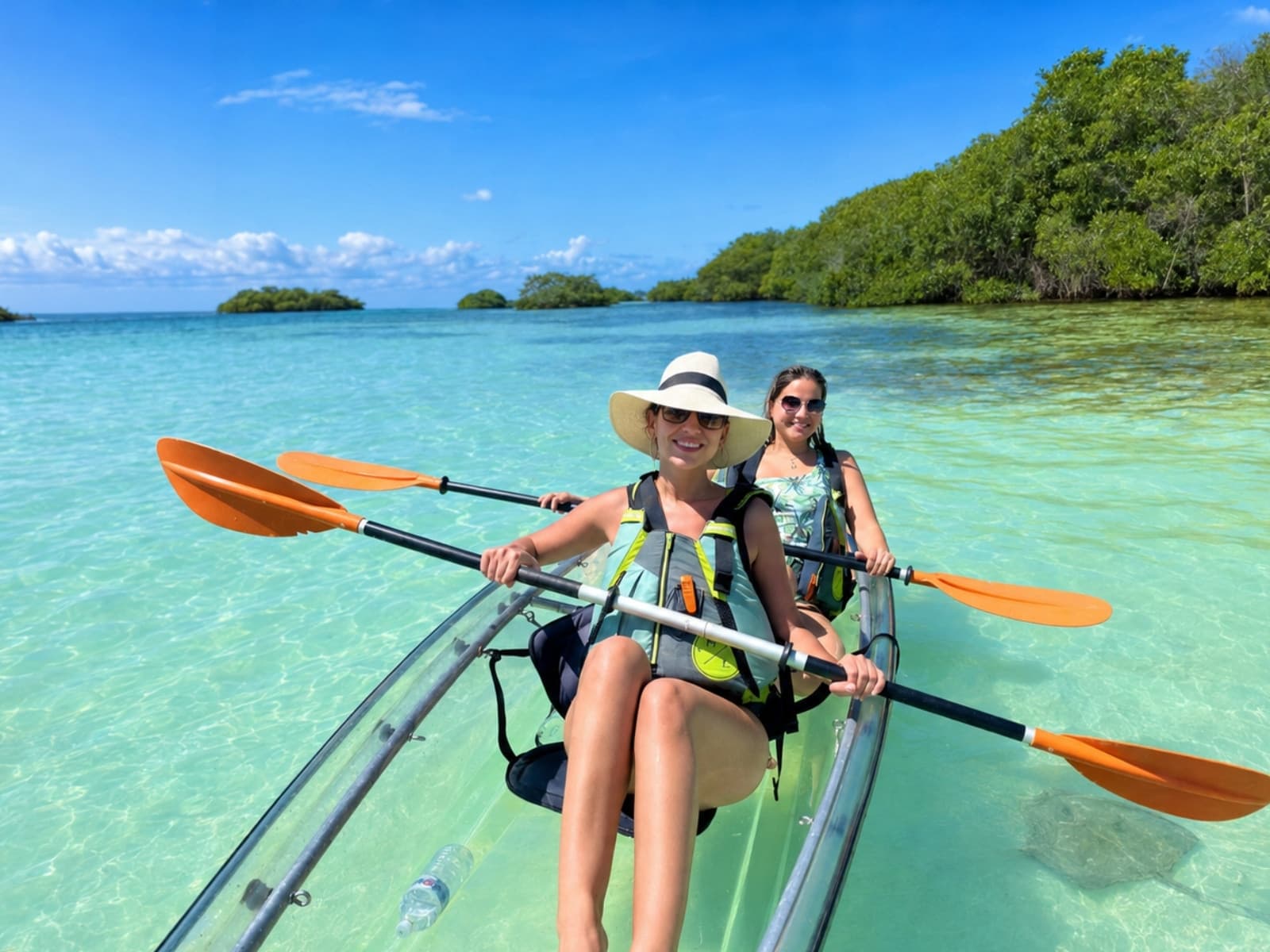 Two women paddle toward the camera in a transparent kayak on turquoise water, with the mangrove behind them