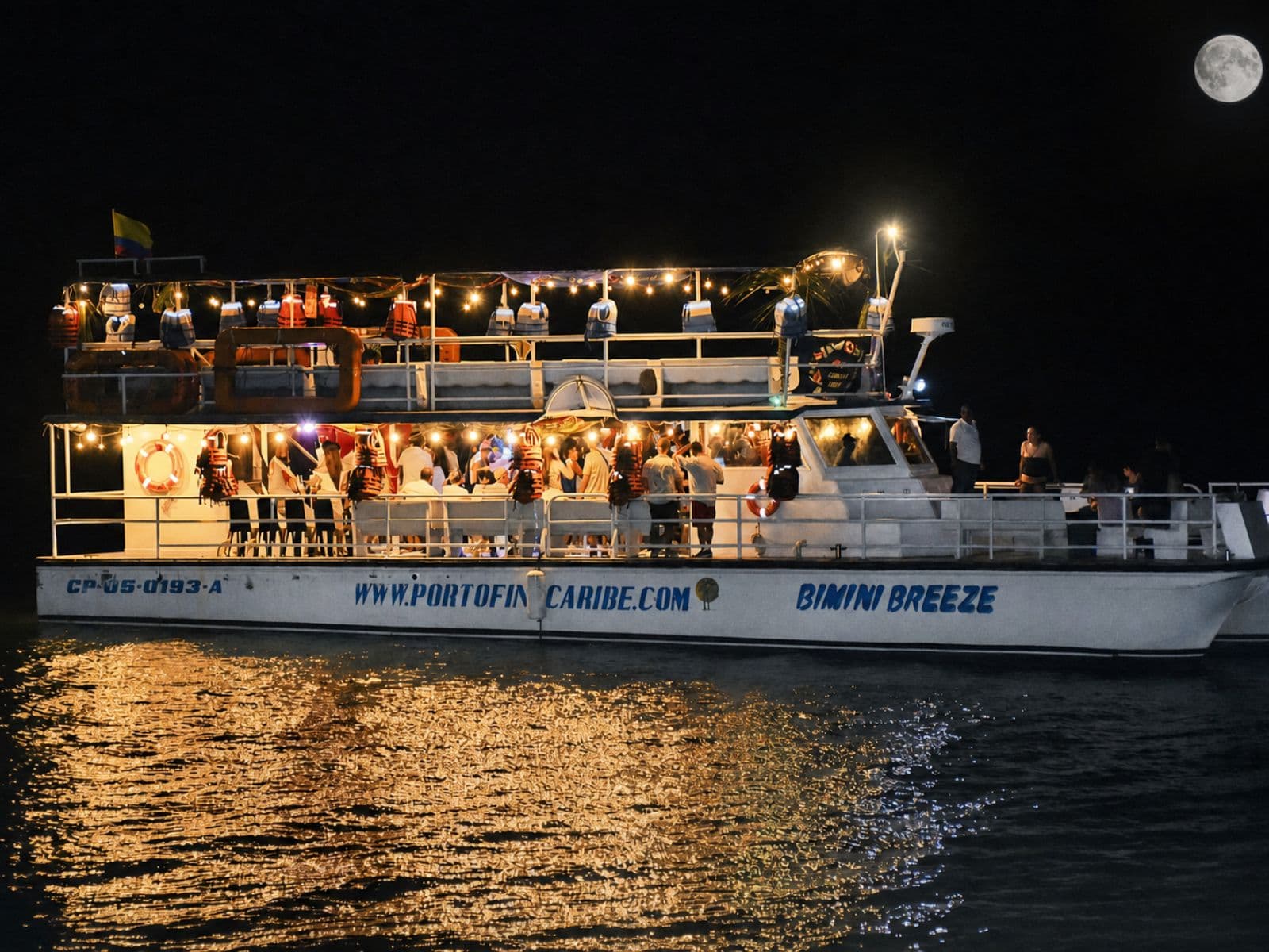 The Bimini Breeze boat, all lit up, sails at night under the moon