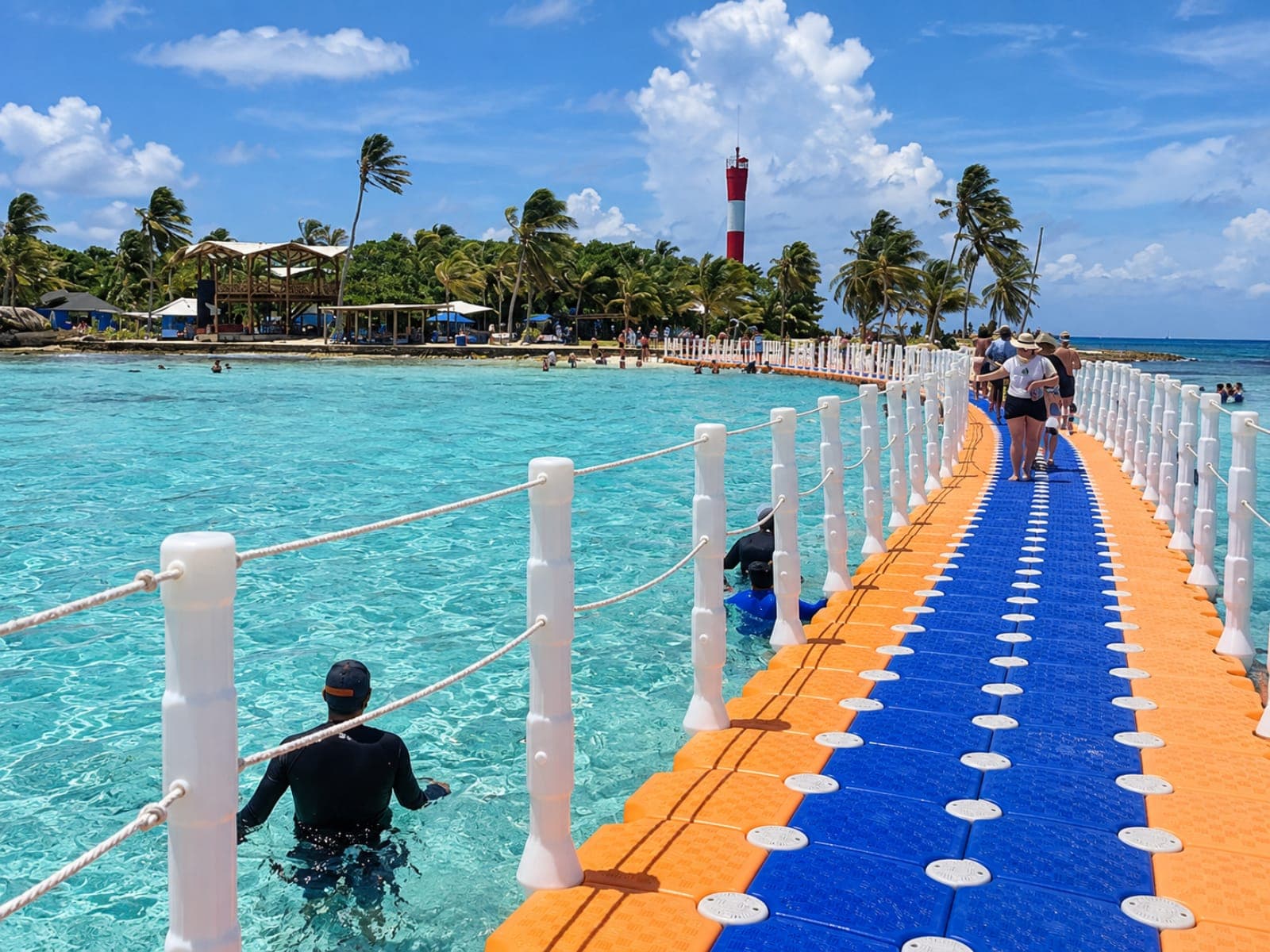 Travelers walking along the colorful floating bridge toward Haynes Cay, with its red and white lighthouse among the palm trees
