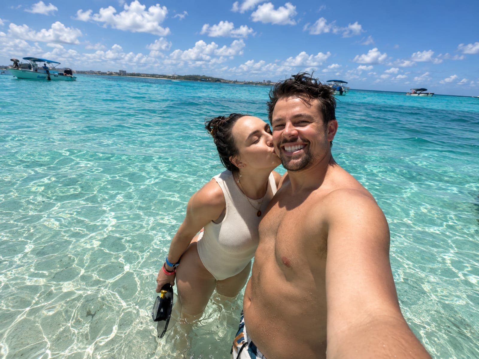 Couple taking a selfie in the turquoise water; in the background, boats and the island