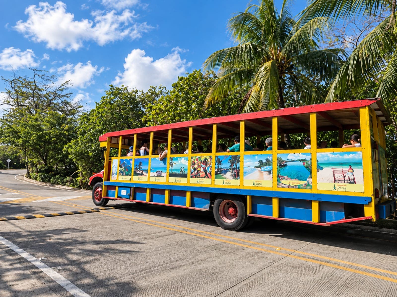 Yellow, red and blue chiva bus with passengers on board and island landscapes painted on its sides, on a road among palm trees