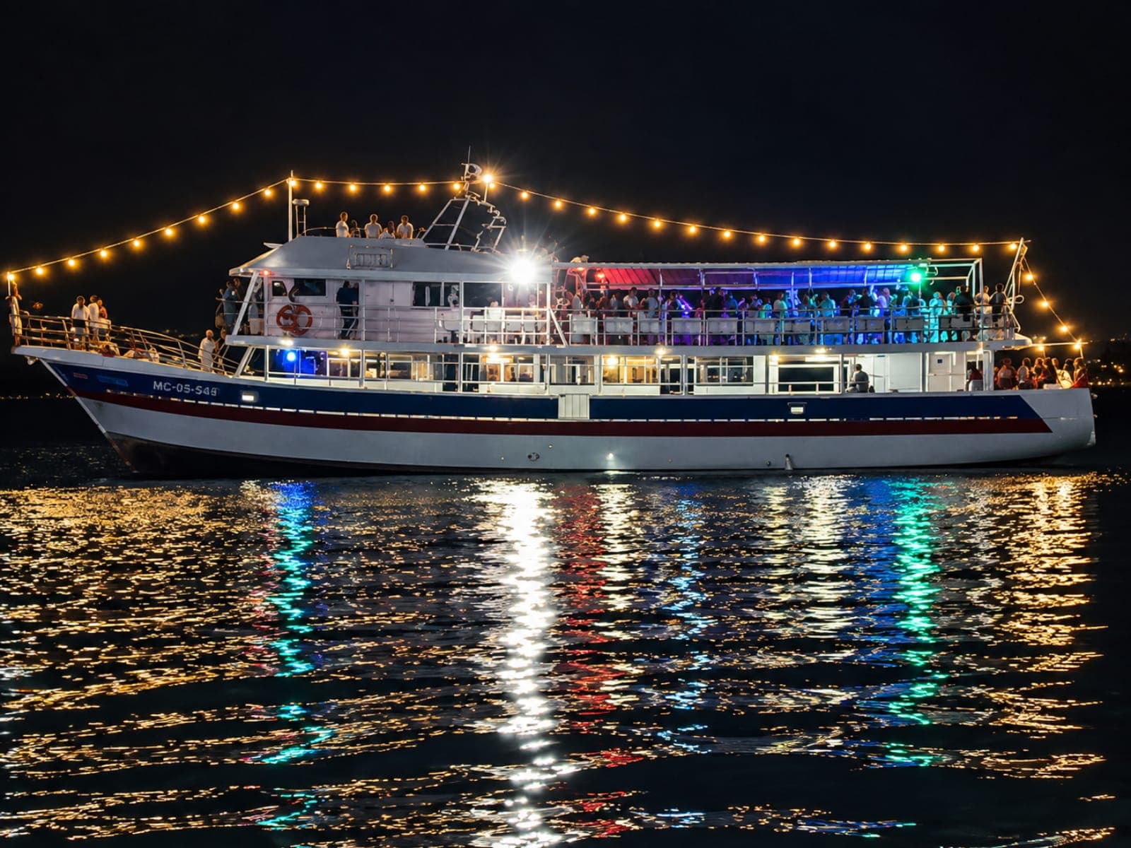 Two-deck yacht lit up with string lights and colored lights, full of travelers, at night on the bay; the lights reflect on the water
